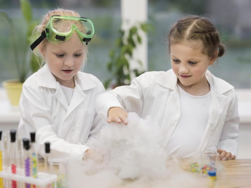 Dwie dziewczynki w białych fartuchach laboratoryjnych przeprowadzają eksperyment naukowy. Jedna ma ochronne okulary, obserwują razem powstającą pianę lub dym z eksperymentu.