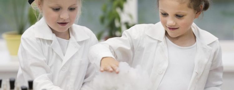 Dwie dziewczynki w białych fartuchach laboratoryjnych przeprowadzają eksperyment naukowy. Jedna ma ochronne okulary, obserwują razem powstającą pianę lub dym z eksperymentu.
