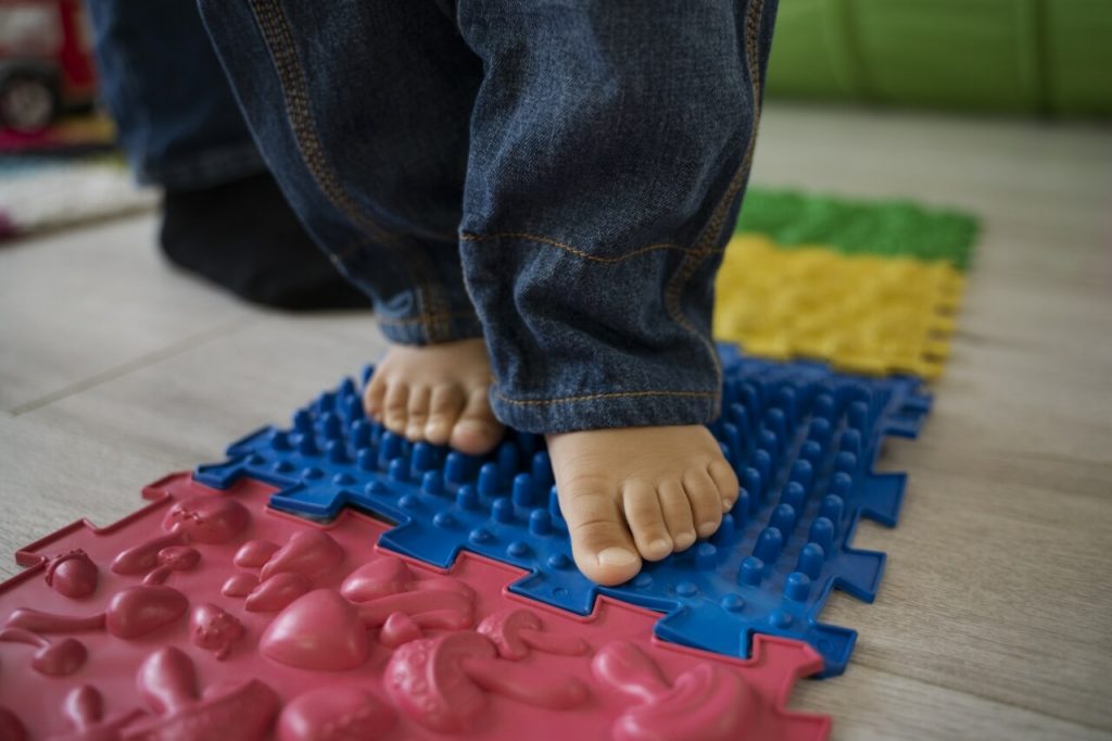 Dziecięce stopy na sensorycznej ścieżce z kolorowych mat o różnych teksturach, w tym niebieskiej z wypustkami i czerwonej z kształtami, wspierających rozwój zmysłów.
