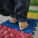 Dziecięce stopy na sensorycznej ścieżce z kolorowych mat o różnych teksturach, w tym niebieskiej z wypustkami i czerwonej z kształtami, wspierających rozwój zmysłów.