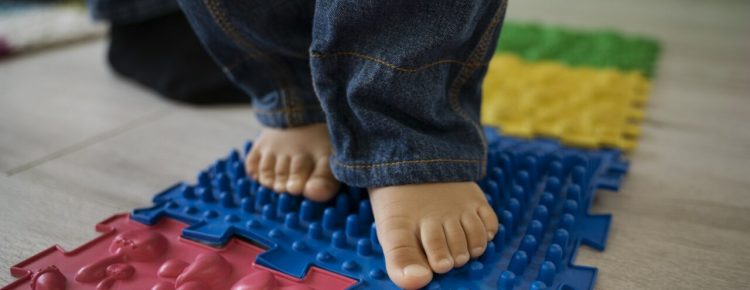 Dziecięce stopy na sensorycznej ścieżce z kolorowych mat o różnych teksturach, w tym niebieskiej z wypustkami i czerwonej z kształtami, wspierających rozwój zmysłów.